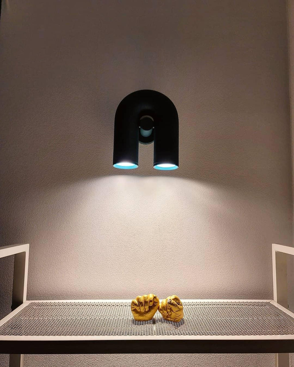 This black Clérachic wall light casts warm downlight over a table, seen from below against a plain wall.