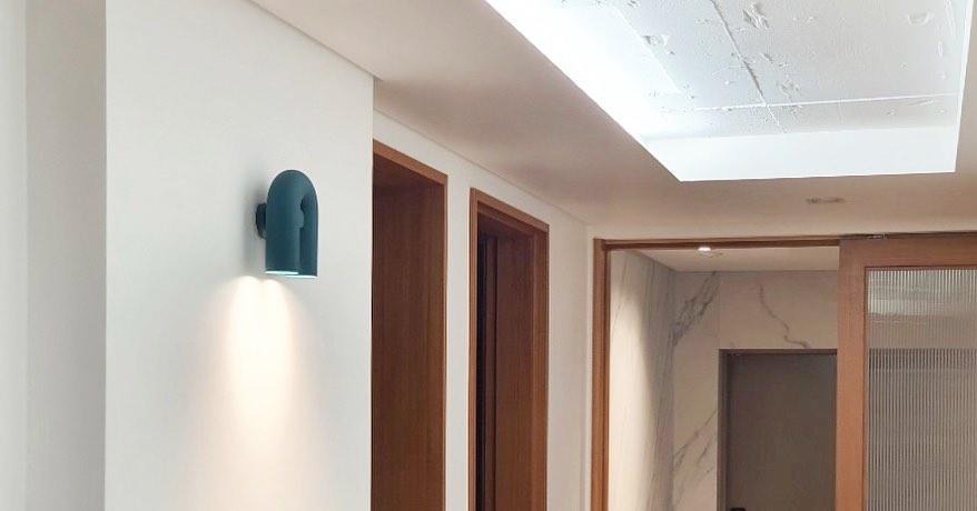 This teal Clérachic wall light glows warm in a skylit hallway lined with wooden doors.