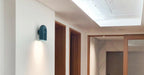 This teal Clérachic wall light glows warm in a skylit hallway lined with wooden doors.