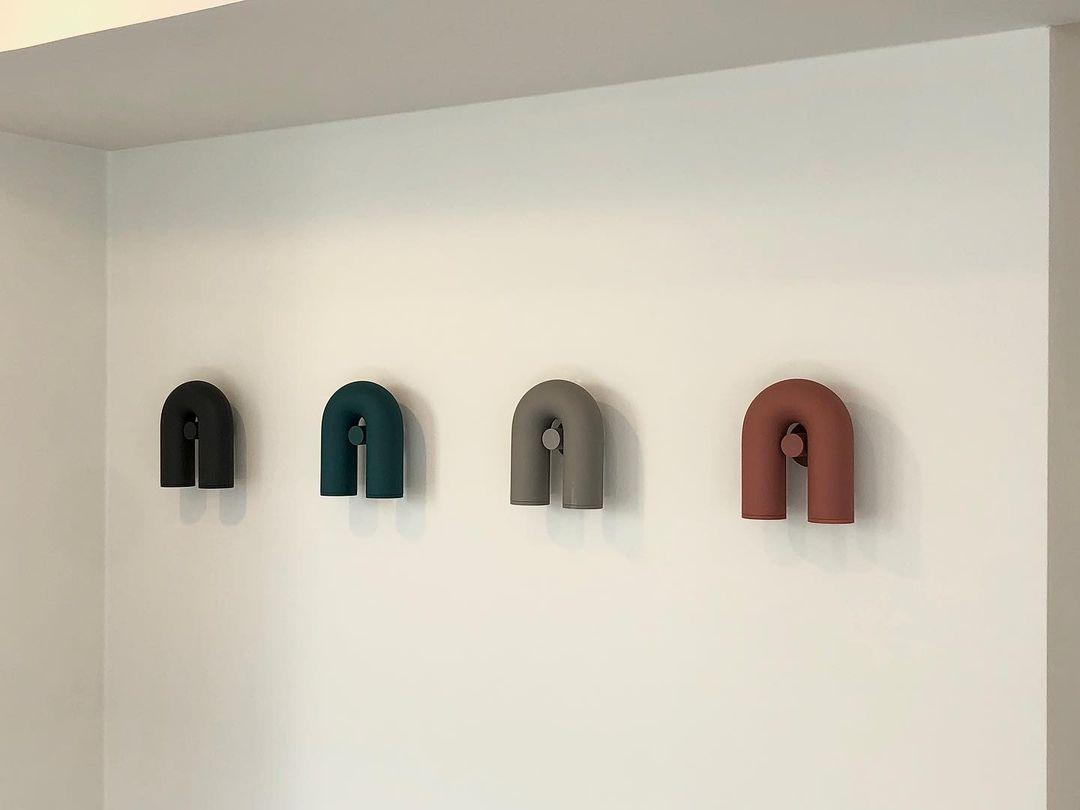 Four Clérachic wall lights in black, teal, grey and terracotta line a plain wall, unlit, for comparison.