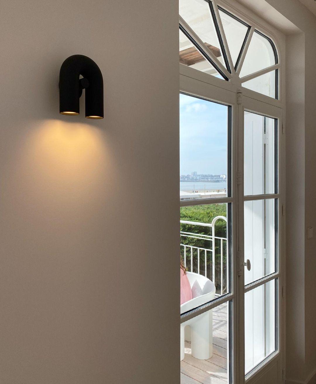 Beside a window overlooking the sea, this black Clérachic wall light glows warm on a plain interior wall.