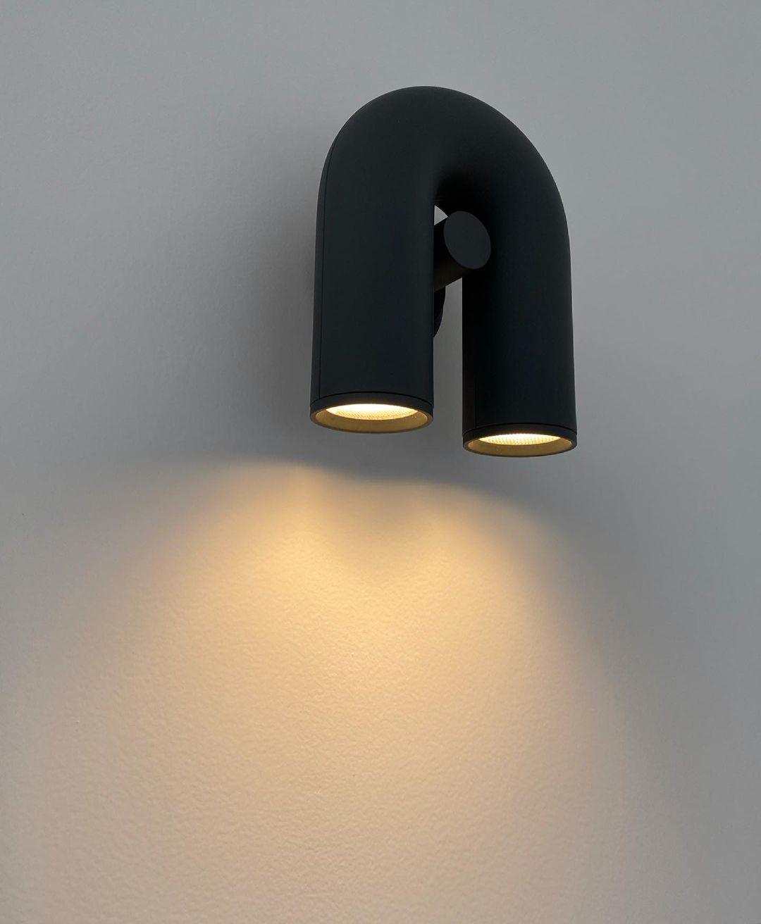 Angled to the side, this black Clérachic wall light glows warm on a plain grey wall.