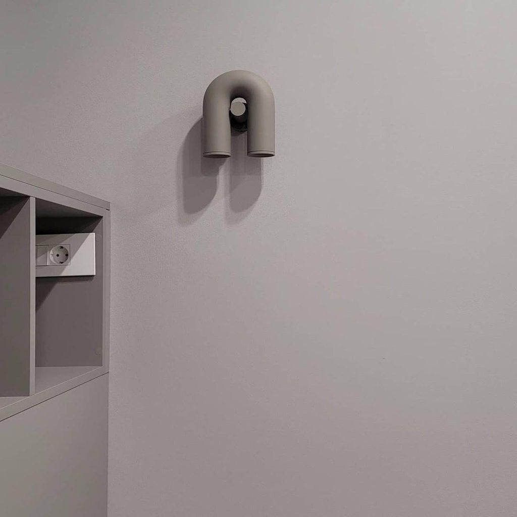 This Clérachic wall light in grey sits small and unlit on a plain white wall beside a wall nook.