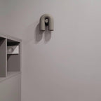 This Clérachic wall light in grey sits small and unlit on a plain white wall beside a wall nook.
