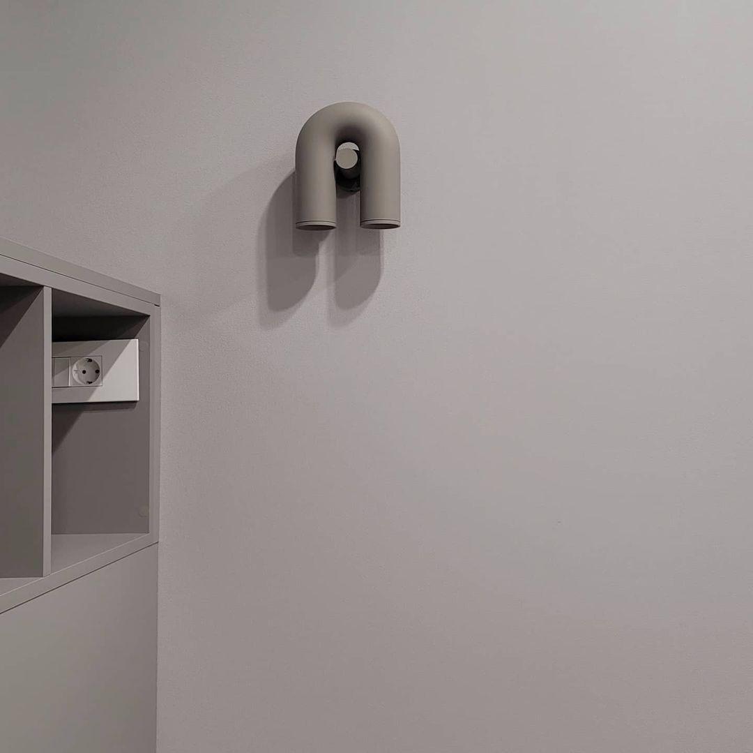 This Clérachic wall light in grey sits small and unlit on a plain white wall beside a wall nook.