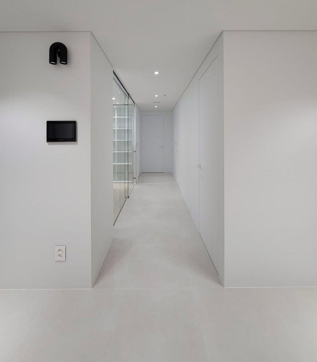 This black Clérachic wall light mounts unlit in a minimalist white corridor, with a wall panel further down the hall.
