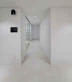 This black Clérachic wall light mounts unlit in a minimalist white corridor, with a wall panel further down the hall.
