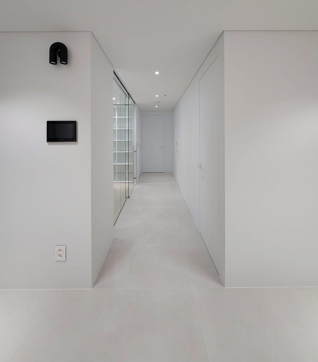 This black Clérachic wall light mounts unlit in a minimalist white corridor, with a wall panel further down the hall.