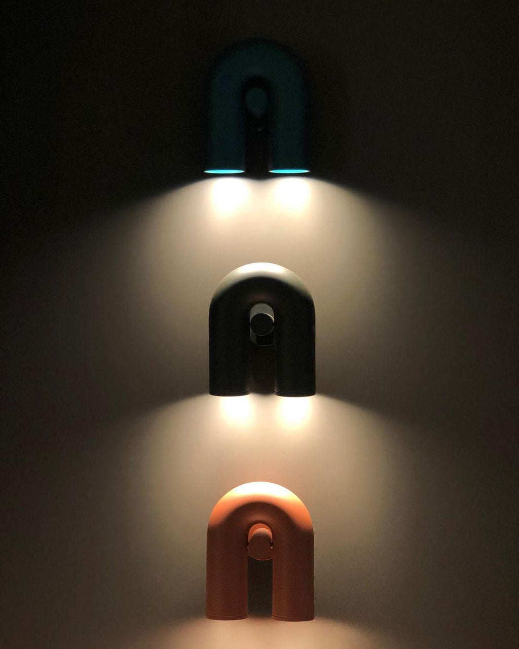 Three Clérachic wall lights in teal, black and terracotta stack down a dark wall, each glowing warm.