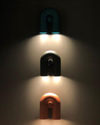Three Clérachic wall lights in teal, black and terracotta stack down a dark wall, each glowing warm.