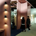 Rows of Clérachic wall lights in teal, black and terracotta climb a pink pillar in a showroom, with visitors walking past in the background.