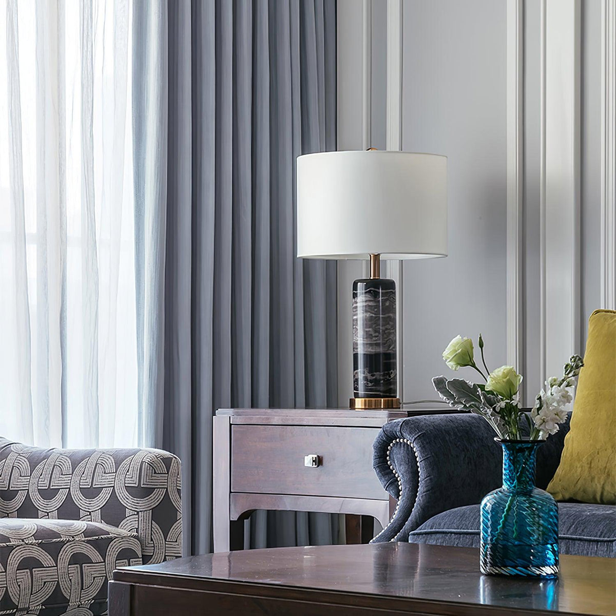 In a room with patterned curtains, the Clérachic table lamp sits beside a blue vase of flowers on a dark table.