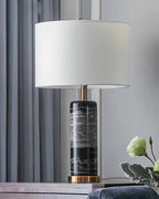 The Clérachic table lamp glows on a tabletop beside a dark curtain and an upholstered ottoman.