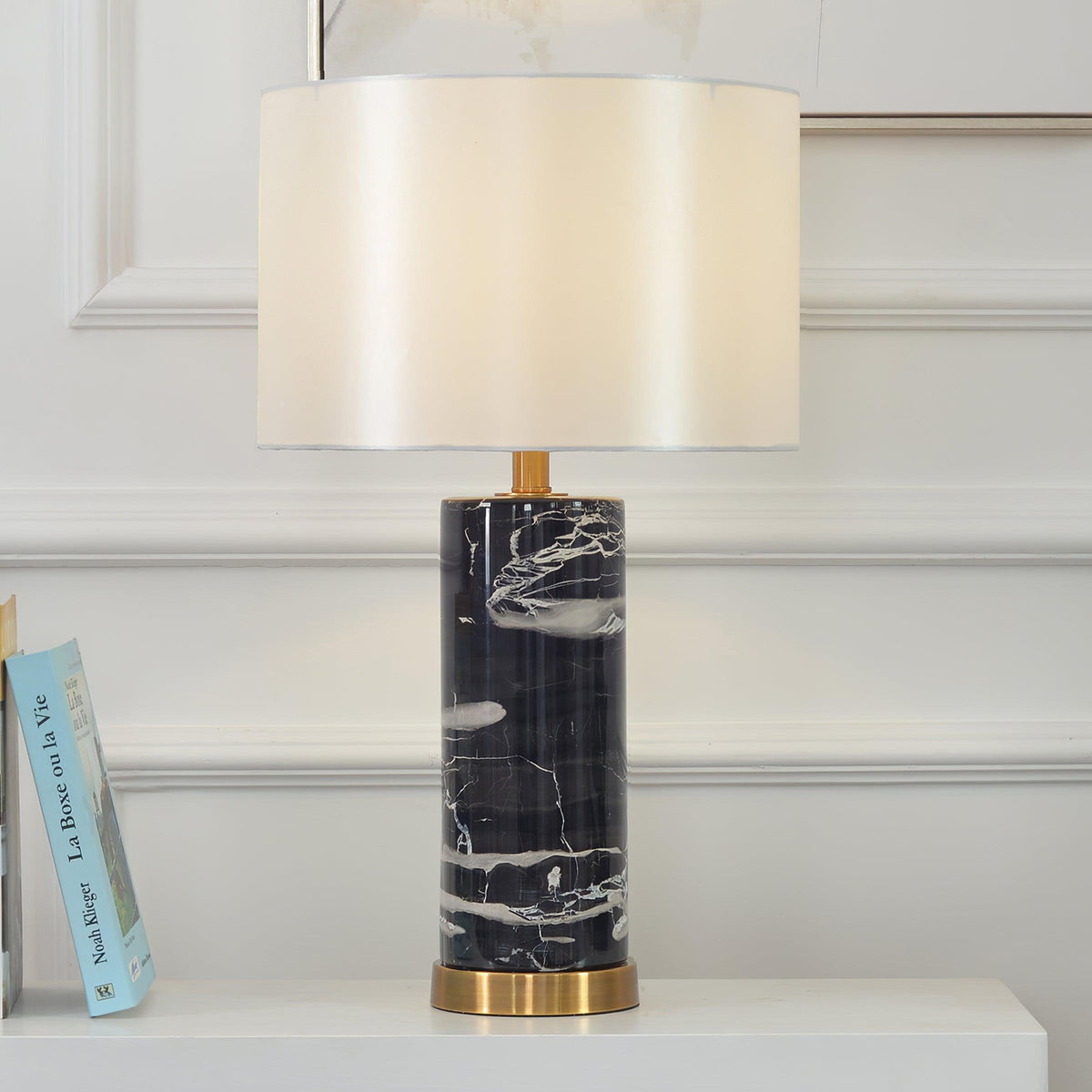 A Clérachic table lamp glows on a bedside surface with books stacked nearby against a white panelled wall.