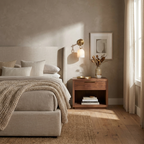 A brass Clérachic wall sconce brightens a sunlit bedroom corner above a walnut nightstand and upholstered bed.