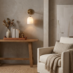 A brass-hardware Clérachic wall sconce lights a console table topped with books and a woven tray in a living room corner.