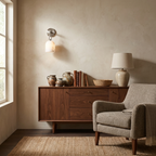 Beside a grey armchair, this brass Clérachic sconce lights a walnut sideboard dressed with pottery, books, and a table lamp.