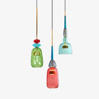 Three Clérachic pendants, green, coral and teal glass shades, hang at staggered heights on a plain white background.