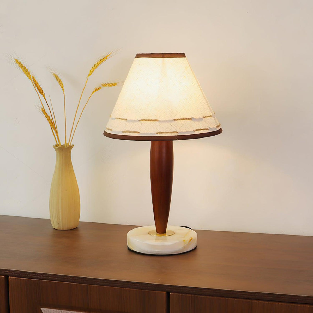Lit Clérachic table lamp beside a dried-wheat vase on a walnut sideboard, its linen shade glowing warm against a cream wall.