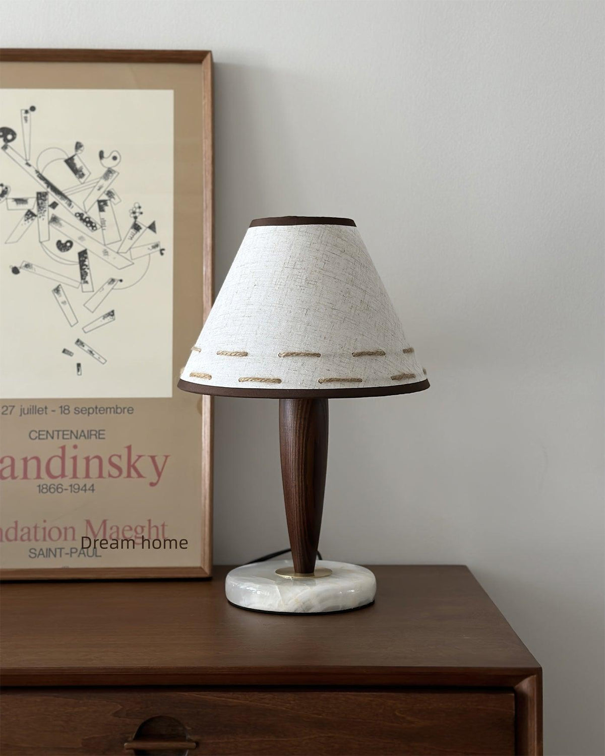 Clérachic lamp lit on a bedside dresser beneath a framed art print, its linen shade casting warm light.