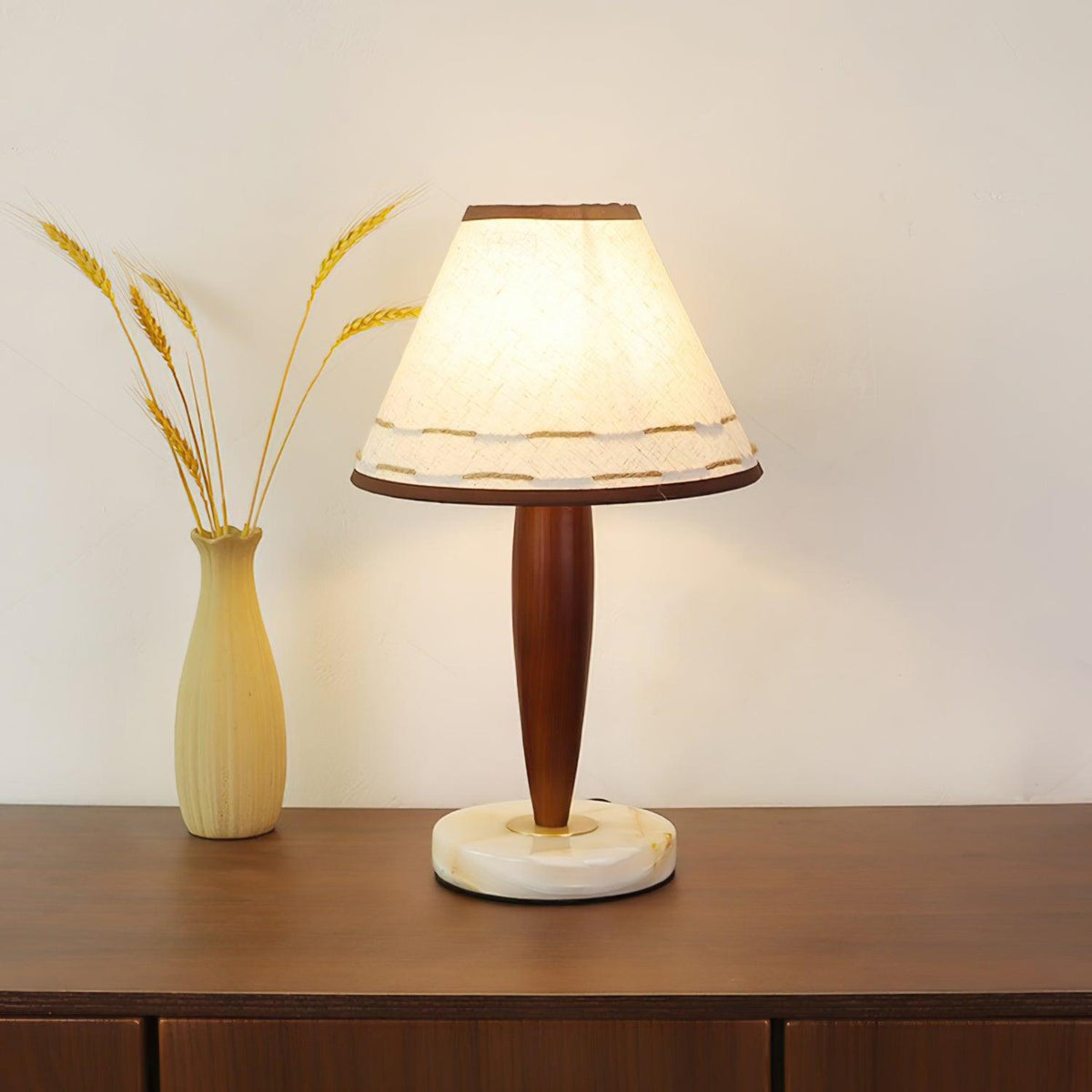 Lit Clérachic lamp beside a dried-wheat vase on a walnut sideboard, cropped closer to show the glowing shade in full.