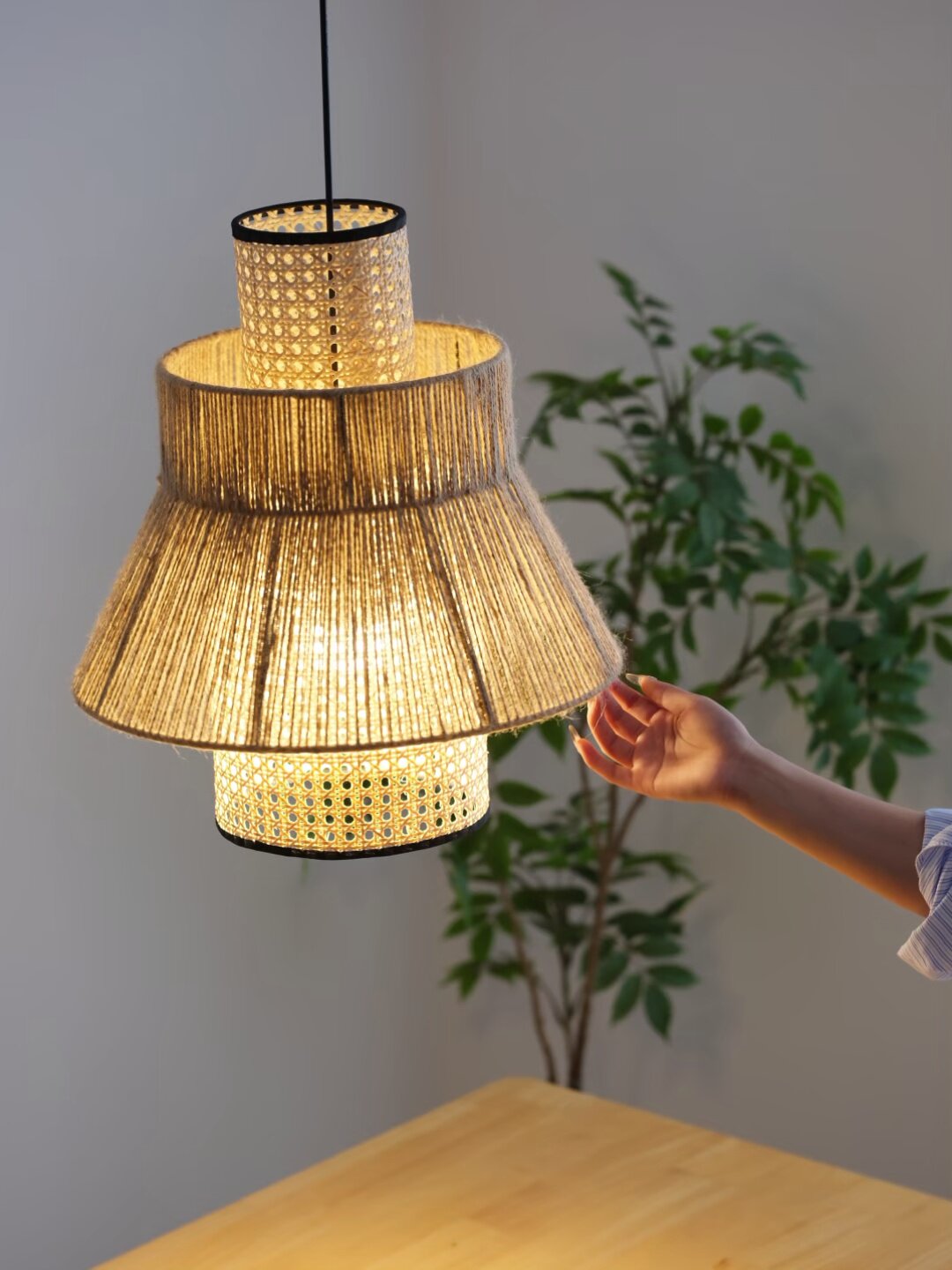 This Clérachic pendant's woven rattan drum and rope fringe glow warm above a wood table beside a potted plant.
