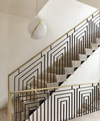 Beside a geometric brass stair rail on a bright landing, a single Clérachic pendant hangs.