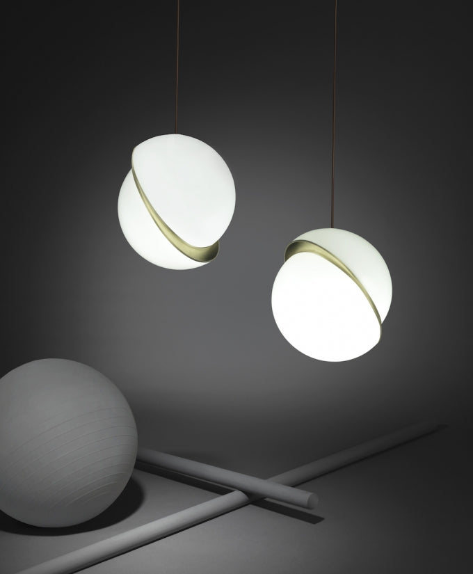 Against a charcoal backdrop, two opal pendants with brass crescents hang beside a grey sphere prop and crossed rods, from Clérachic.