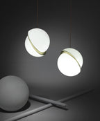 Against a charcoal backdrop, two opal pendants with brass crescents hang beside a grey sphere prop and crossed rods, from Clérachic.