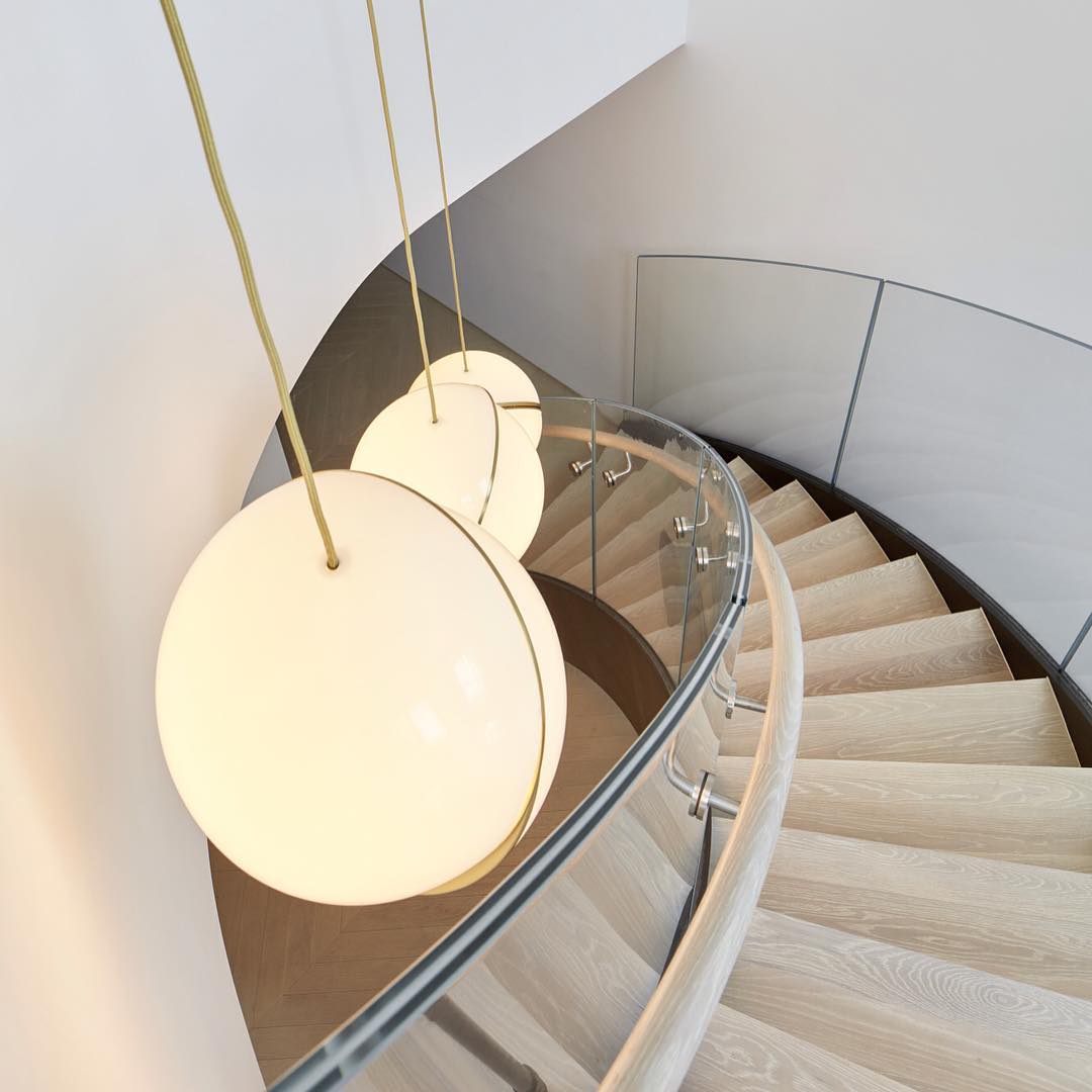 Beside a curved staircase with a glass balustrade, clustered Clérachic pendants glow, seen close up.