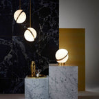 Beside dark marble plinths and a brass block, two opal spheres with brass crescents hang, from Clérachic.