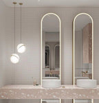 Beside arched mirrors above a pink terrazzo vanity, two pendants of different heights hang, from Clérachic.