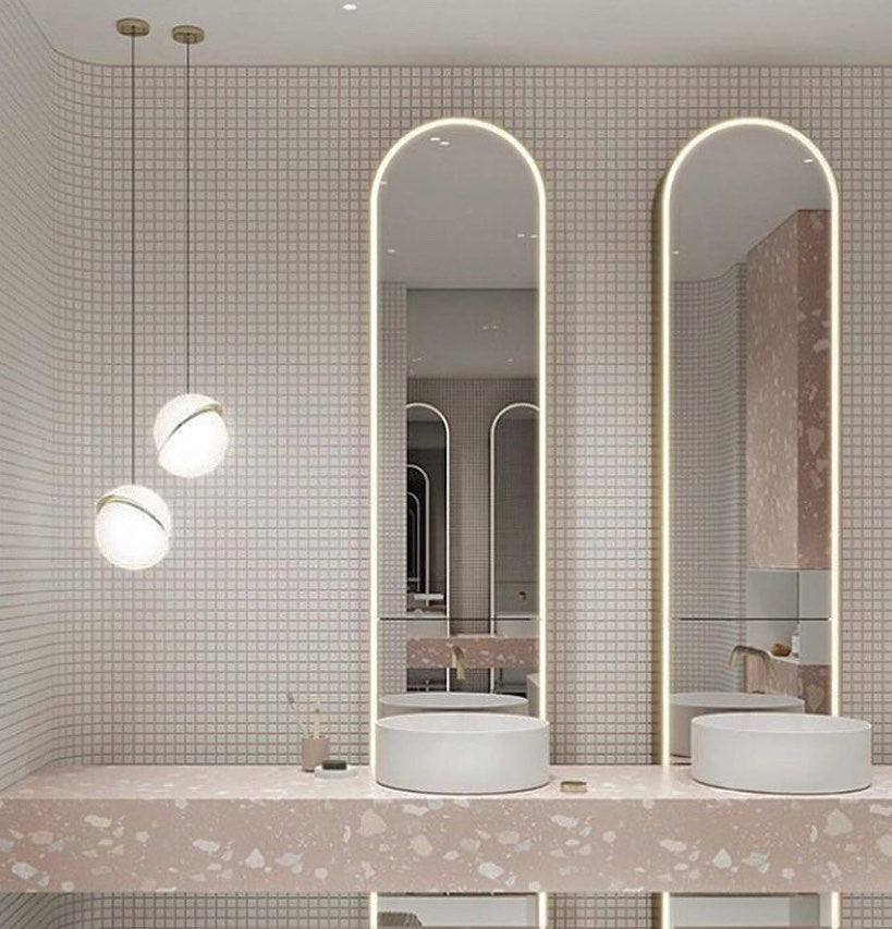 Beside arched mirrors above a pink terrazzo vanity, two pendants of different heights hang, from Clérachic.