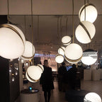 Above a lit shopfloor with visitors browsing nearby, many opal spheres with brass crescents cluster, from Clérachic.