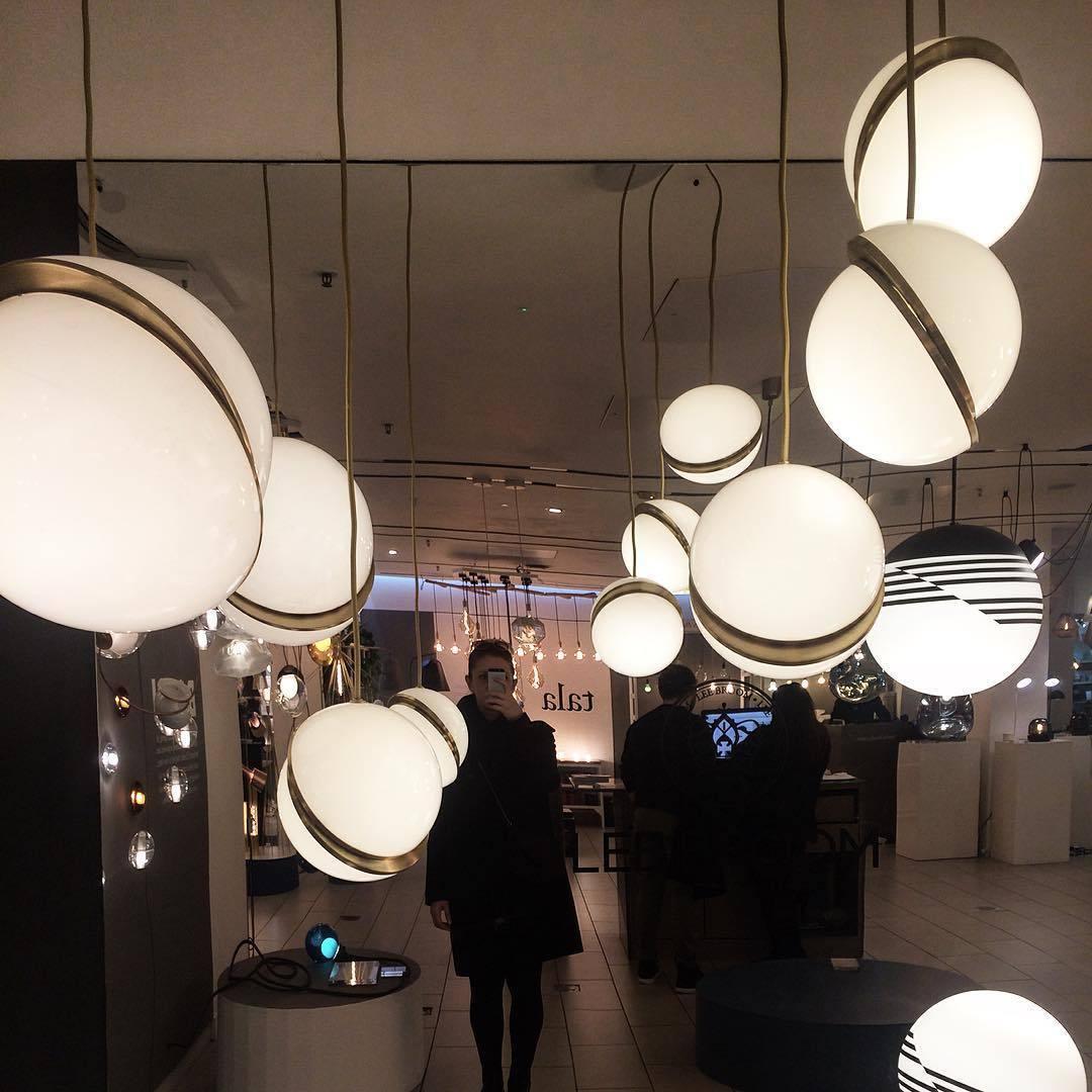 Above a lit shopfloor with visitors browsing nearby, many opal spheres with brass crescents cluster, from Clérachic.