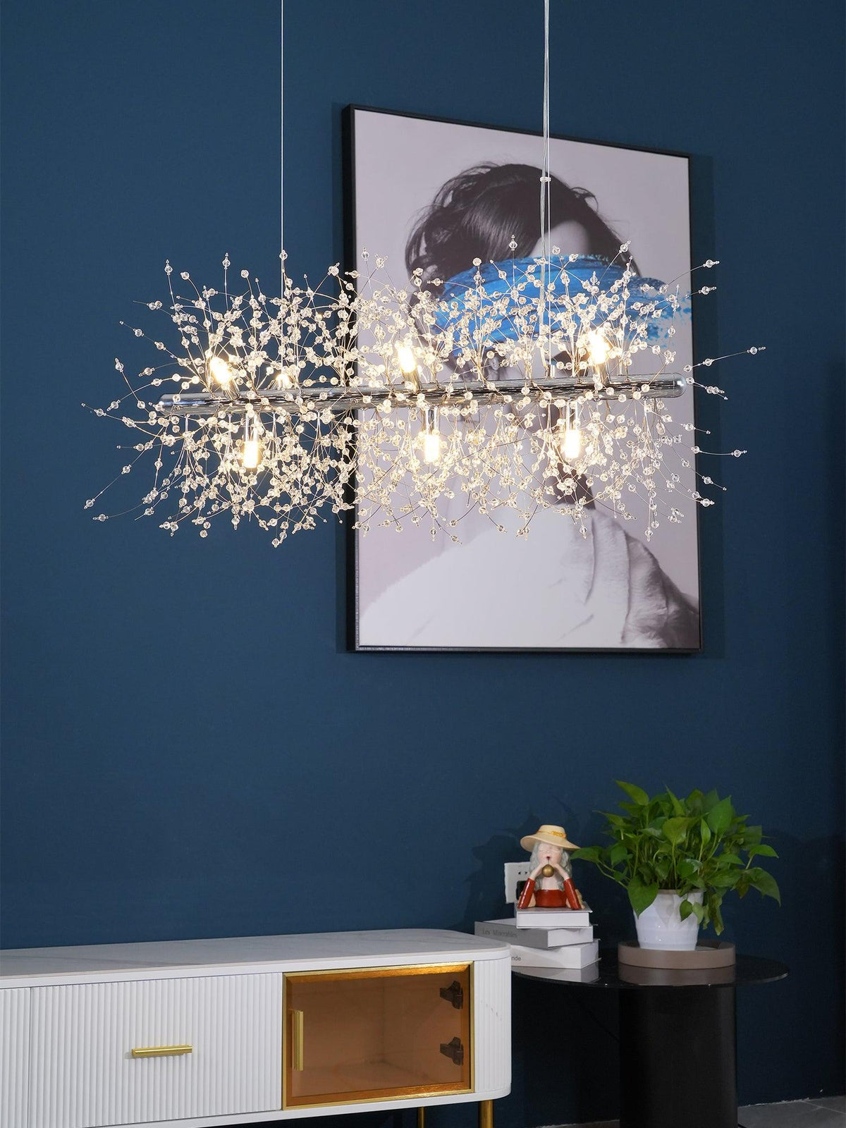 This Clérachic chandelier hangs lit against a navy wall beside a framed portrait print and a console table with a plant.