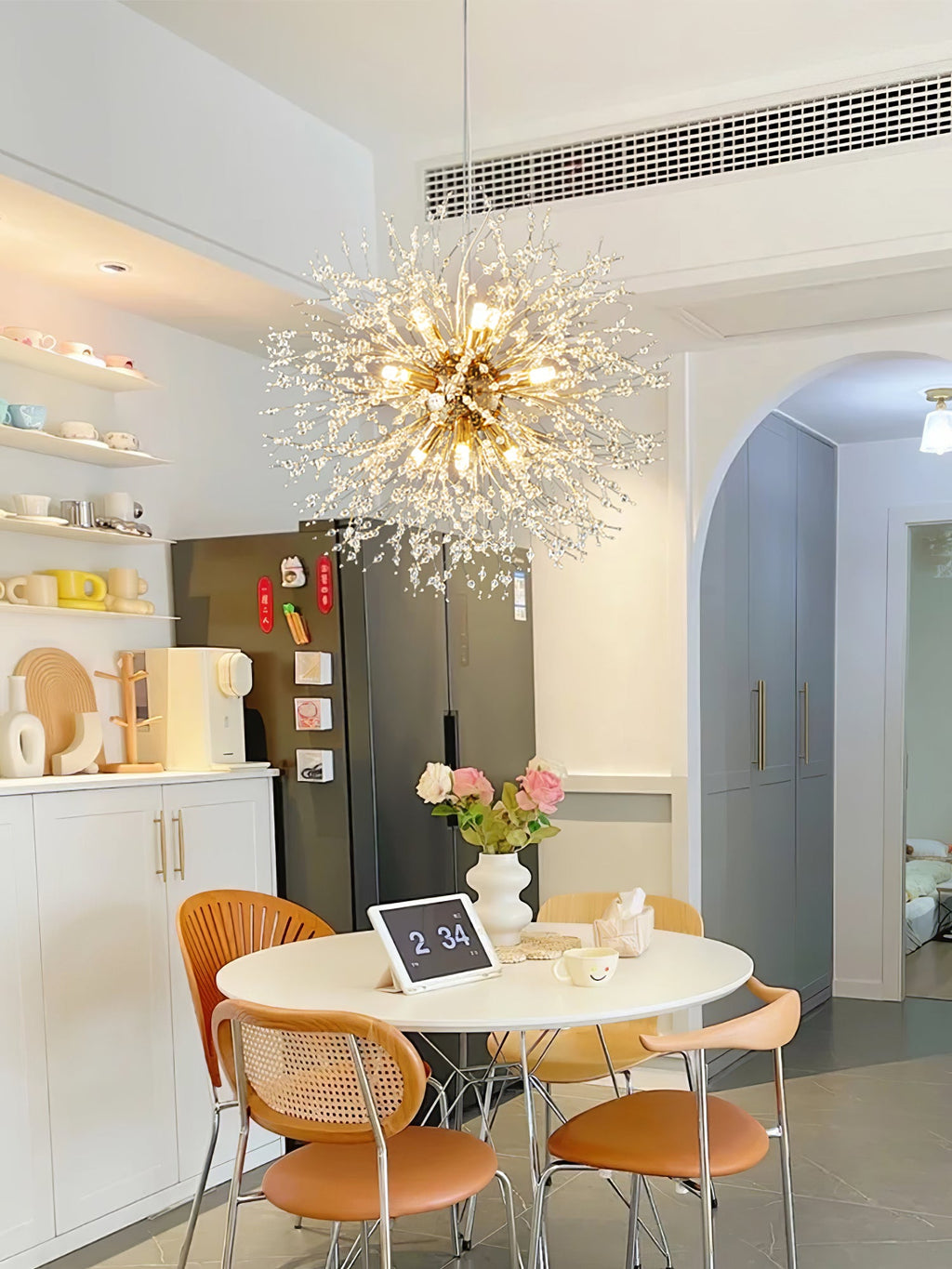 Another view of this Clérachic chandelier lit above a round dining nook with open shelving and a stainless fridge.