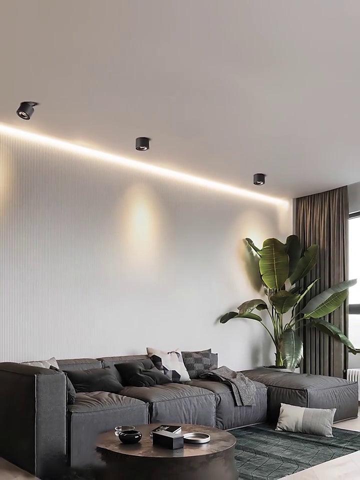 Black units of this Clérachic downlight line a recessed wall channel above a dark sofa and a large potted plant.