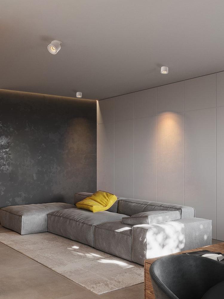 A black unit of this Clérachic downlight is mounted in a lounge corner above a grey sectional sofa and yellow cushion.