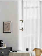A single wide-cylinder Clérachic pendant hangs near a window beside a dark sideboard and a rattan chair.