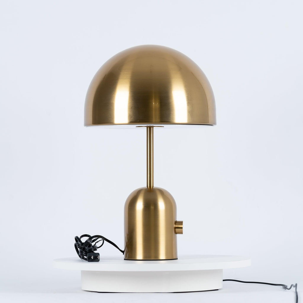 This Clérachic table lamp in a brushed gold finish stands on a white pedestal, its cord trailing beside it.