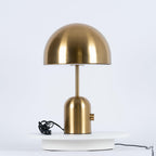 This Clérachic table lamp in a brushed gold finish stands on a white pedestal, its cord trailing beside it.