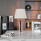 A Clérachic table lamp sits unlit on a stone plinth, its shade reading pale grey against the dark wood shelving.