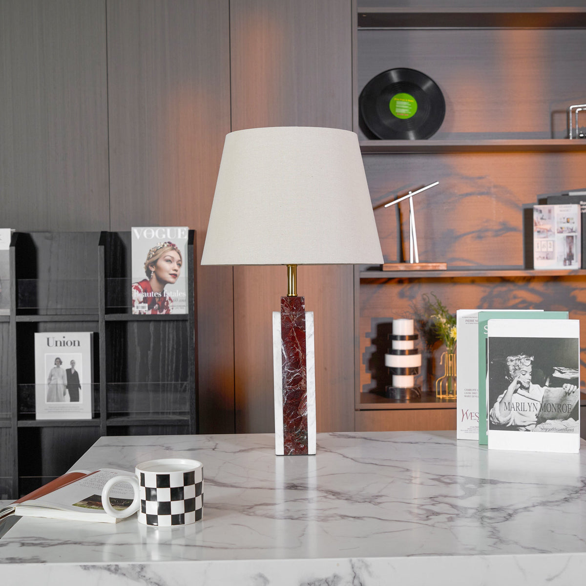 A Clérachic table lamp sits unlit on a stone plinth, its shade reading pale grey against the dark wood shelving.