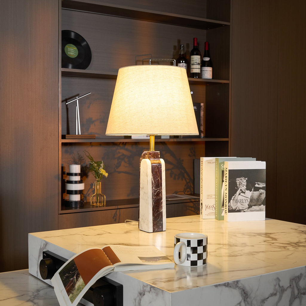 A Clérachic marble table lamp lit on a white stone table, a magazine draped off the edge beside books and a mug.