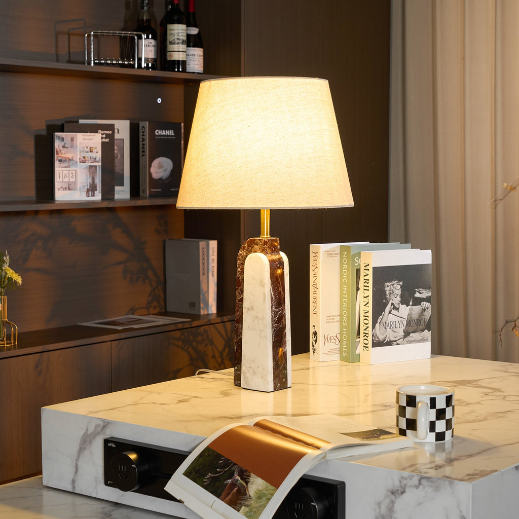 A Clérachic marble table lamp on a white stone side table, styled with stacked books, a checkerboard mug and a magazine.