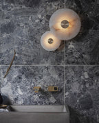 Two alabaster disc sconces lit above a concrete basin and brass tap on a grey marble bathroom wall.