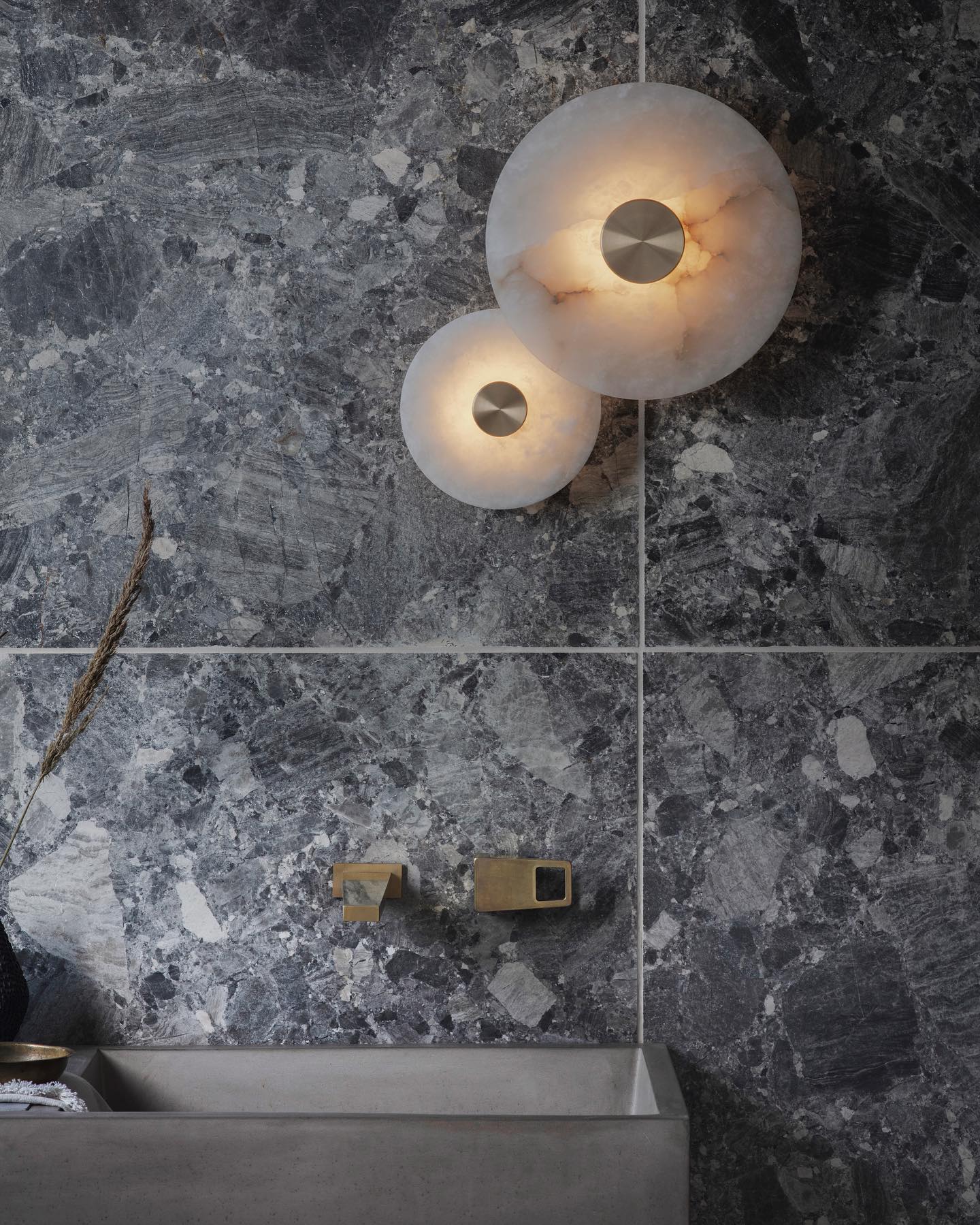 Two alabaster disc sconces lit above a concrete basin and brass tap on a grey marble bathroom wall.