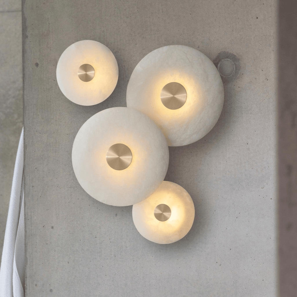 Four alabaster disc wall lights in graduated sizes, lit and clustered on a concrete wall.