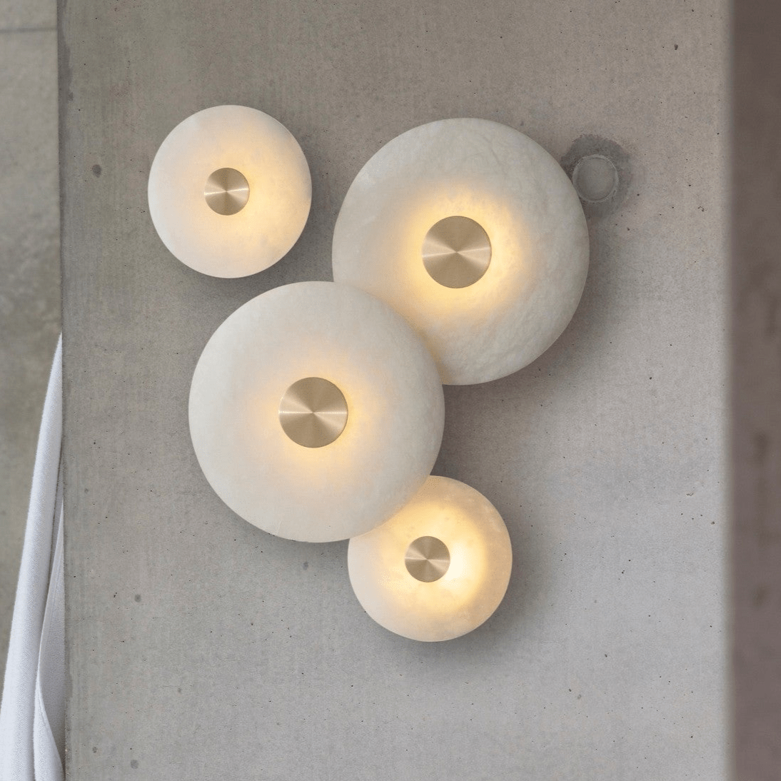 Four sizes of the alabaster disc wall light arranged as one lit composition on bare concrete.